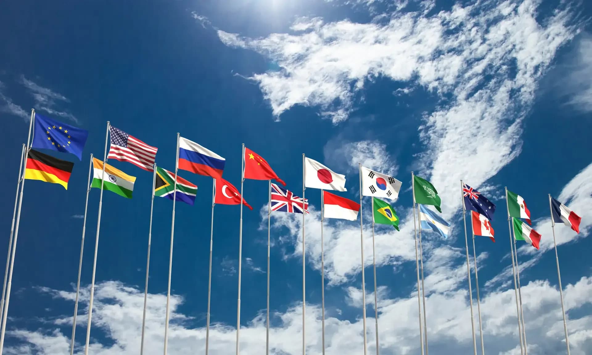 Vários mastros enfileirados exibindo bandeiras de diversos países, como Brasil, EUA, China e Reino Unido, sob um céu azul com nuvens esparsas